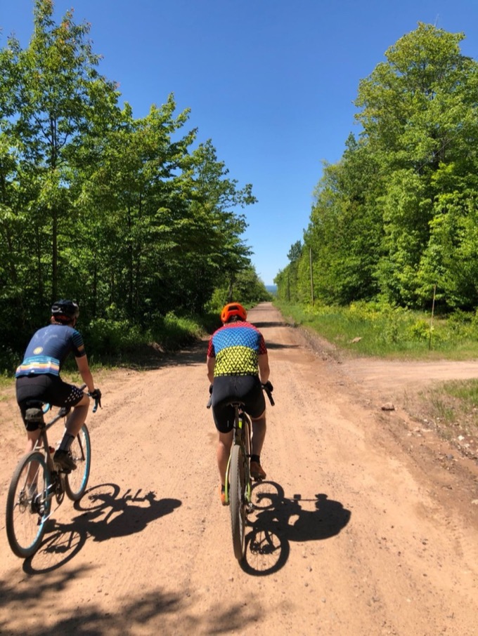 Two riders on a wide sandy road lined with trees
